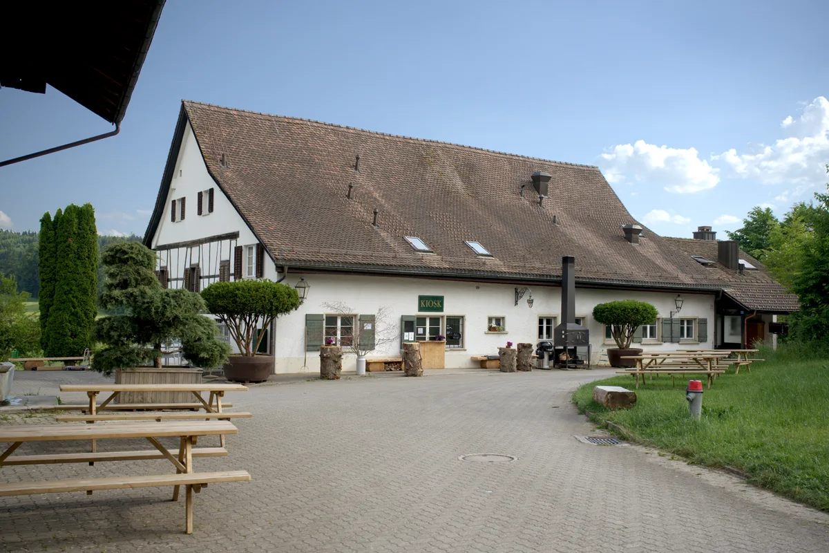Die Bänke und Tische geben eine erste Vorstellung, wie gemütlich es einst sein wird, bei schönem Wetter ein kühles Getränk zu schlürfen. Ein Haus und ein Vorplatz sind zu sehen.